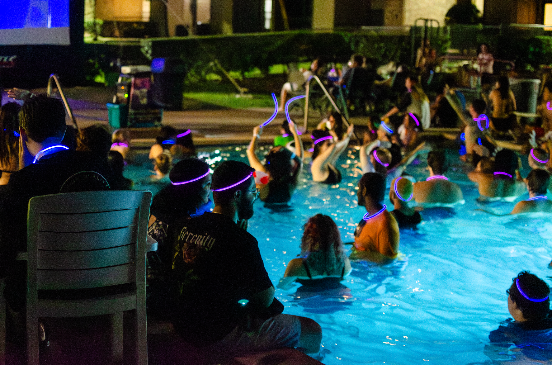 A variety of people, some with glow sticks, lounge in a pool at night.  
